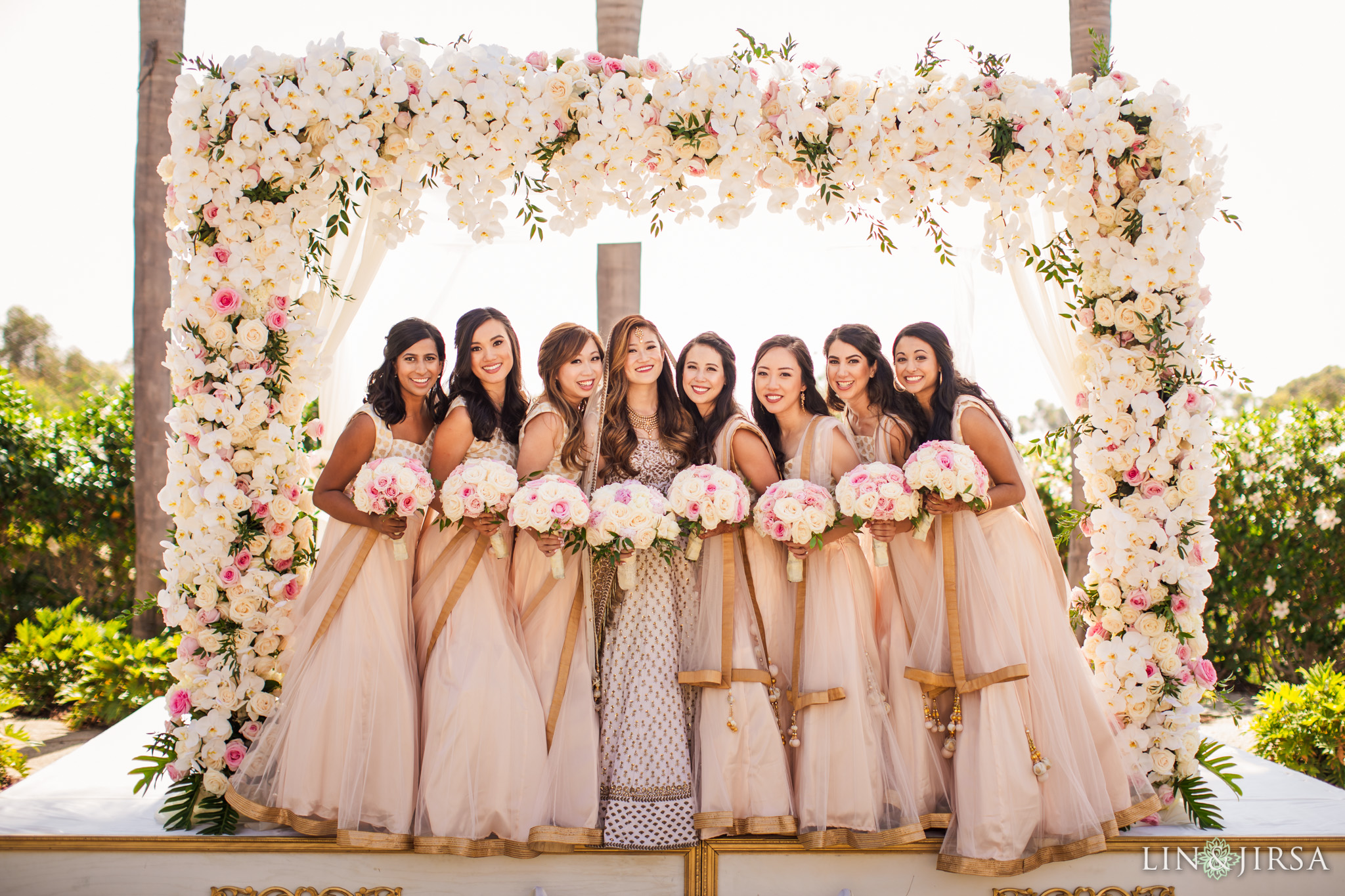 Seven bridesmaids posing with white and pink rose flower bouquets in their hands, with the bride, under a white rose archway and palm trees, near the beach for Lin and Jirsa Los Angeles at the Park Hyatt Aviara Resort in Carlsbad, California