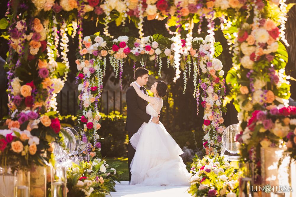 A wedding ceremony photograph shot by Lin and Jirsa Los Angeles with the bride and groom holding each other under ornate wedding arches that are draped with orange, pink, yellow and red roses and wisteria flowers, planned by Christina of Lavish Weddings at the San Diego Museum of Art , in San Diego, California